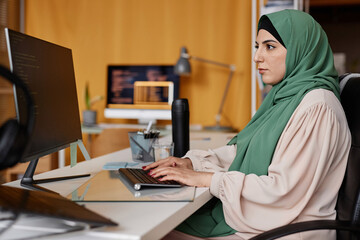 Side view portrait of successful Muslim woman as female programmer writing code in office and wearing modest clothing