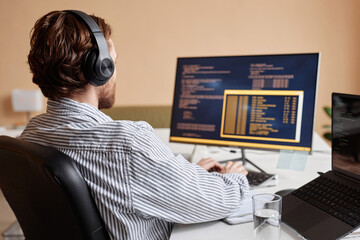 Male computer programmer writing code at office workplace and wearing headphones, copy space