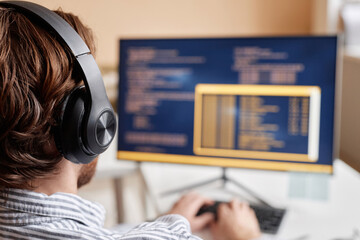 Over shoulder of male computer programmer writing code in office and wearing big headphones, copy...