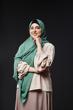 Vertical portrait of young Muslim woman wearing modest clothing and smiling at camera standing against black background in studio