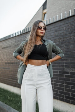 Chic street beauty model girl with vintage sunglasses in fashion clothes with a shirt stands near a black brick fence