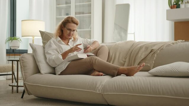 Pretty mature blonde in glasses in white T-shirt and beige trousers having rest on cozy sofa, holding pencil, reading thick book in a home atmosphere. High quality 4k footage