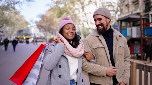 Cheerful multiracial couple walking in the city doing christmas shoppings together, holding gift bags, pointing at things and smiling, having a good time outdoors in winter season.
