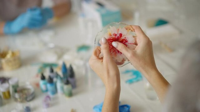 Close-up female hands holding beautiful craft made of transparent epoxy art resin with blooming red flower inside on white background. Working process in workshop. Hobbies and creativity concept