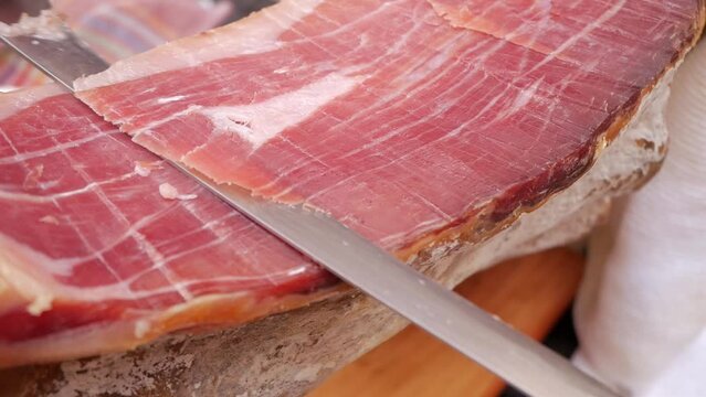 Closeup shot, cured ham, spanish serrano ham, being sliced for tapas meal. Large thin sharp blade knife slicing red meat with fat traces lines across.