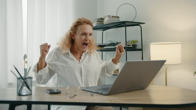 Attractive neat woman with short curly blode hair sitting at table in cozy room with light interior, typing on laptop, clenching fists with excitement. High quality 4k footage