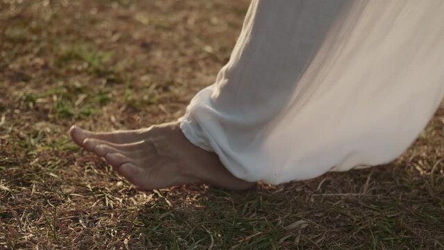 Barefoot woman in white transparent wide trousers practices qigong on the ground. Foot stretch. Interaction of matter, energy and spirit. 
