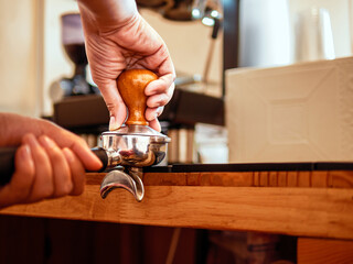 Hand of a barista holding a portafilter and a coffee tamper making an espresso coffee. Barista presses ground coffee using a tamper in a coffee shop
