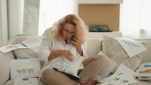 Blonde woman in glasses sitting in yoga pose on white sofa at home reviewing documents with irritation, talking on cell phone, emotionally waving hands. High quality 4k footage
