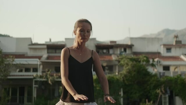 A European woman in a black sleeveless tank top practices breathing and movement exercises with her eyes closed outdoors. Body position control. Qigong. Tai Ji. Chinese traditional exercises.