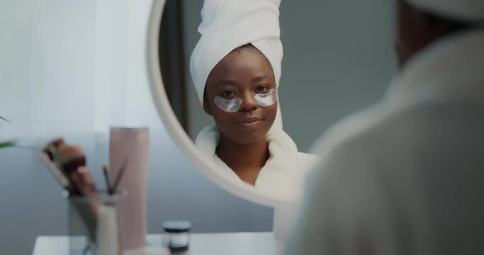 African american woman in bathrobe and towel on head applying eye patches.