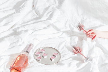 Woman hand holding shiny champagne glass, bottle of sparkling rose wine, pink candle hearts on bed cloth. Minimal lifestyle aesthetic photo, romance mood, Valentine's Day, romantic love concept