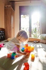 Little girl builds a house from Lego blocks while squatting on the bed. Side view