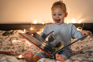 Little girl reading pop up book in home bed in Christmas environment with lights background