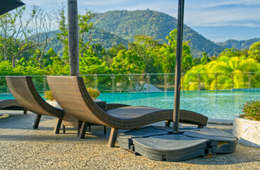 Empty two wicker sun beds beside a swimming pool of a resort, morning light, background of trees, mountain, and sky.