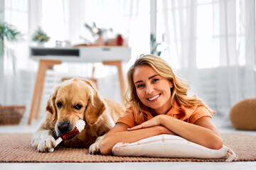 Leisure with pets. Portrait of a beautiful blonde woman and her dog. A golden retriever chews on a toy in the shape of a chicken leg and looks at the camera. - obrazy, fototapety, plakaty