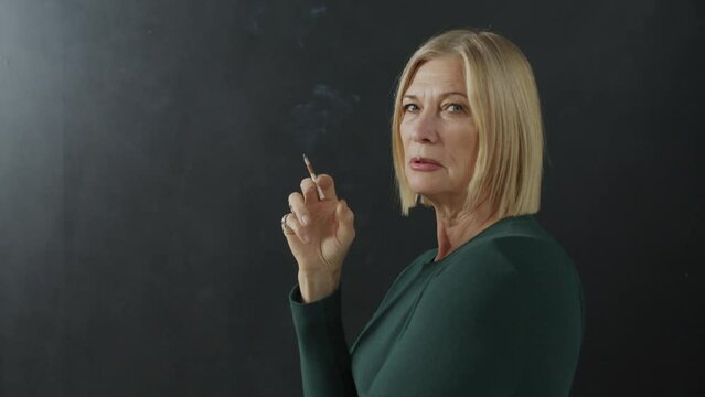 Studio shot of elegant senior Caucasian woman wearing dark green clothes smoking cigarette, black background
