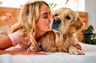 Sincere love to pet. Friendly young woman kissing muzzle of golden retriever while lying together on soft bed. Female blonde strengthening friendship bond with canine buddy at home. - obrazy, fototapety, plakaty