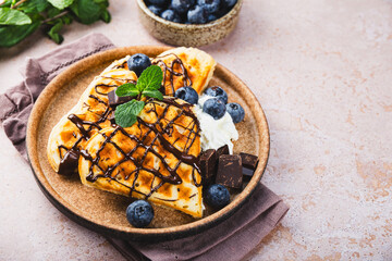 Homemade waffles with blueberries, chocolate sauce and cream cheese in a plate and a beige background