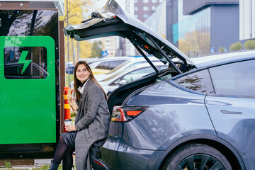 Young woman waiting for her electric car charging, sitting in a car trunk and drinking coffee....