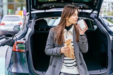 Charming young woman sitting in the car trunk with coffee while recharging electric vehicle with...