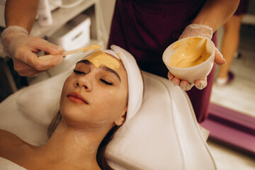 Cosmetic jelly mask, facial skincare. Cosmetologist applies yellow gold alginate mask with spatula...