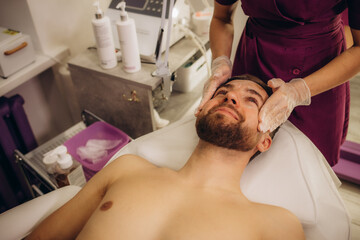 Obraz na płótnie Canvas Beautician applying scrub onto young man's face in spa salon
