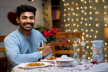 Fototapeta na wymiar Happy smiling indian young man with red roses looking at camera during candlelight dinner at home - concept of enjoyment, valentines day preparation and dating