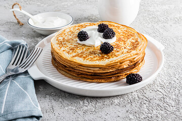 A large stack of delicious hot pancakes with sour cream and blackberries on a beautiful white serving tray with cutlery and a plot. Front view.
