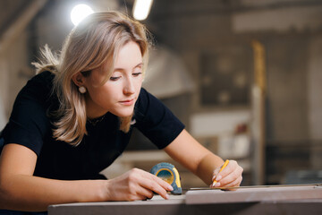 Young woman worker marks with pencil dimensions of wooden furniture for cutting on machine. DIY...