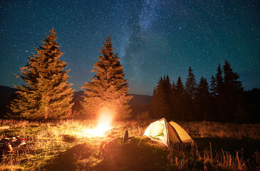 Night camping in mountains under starry sky and Milky way. Female tourist resting near burning campfire and illuminated tent in campsite, lying on grass and enjoying night sky with stars.