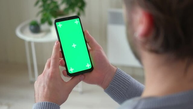 Young man sitting at home holding smartphone green mock-up screen in hand. Male person using chroma key mobile phone. Vertical mode. Touching, swiping display, tapping, surfing Internet social media	
