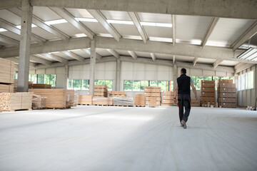 A worker leisurely strolls through a warehouse housing a diverse array of wooden products,...