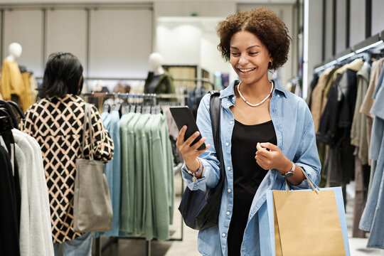 Laughing Young African American Woman Looking At Smartphone Screen While Shopping At Clothing Store