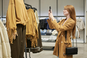 Medium full shot of young caucasian woman taking picture of outfit put on mannequin