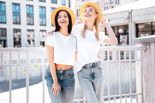 Two young beautiful smiling hipster female in trendy summer white t-shirt and jeans clothes. Carefree women posing in street. Positive models having fun. Cheerful and happy. In hat