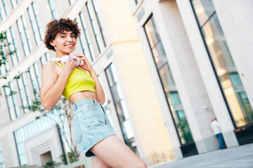 Young beautiful smiling hipster woman in trendy summer clothes. Carefree woman with curls hairstyle, posing in the street at sunny day. Positive model outdoors. Listens music at her headphones on neck