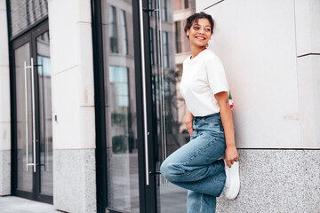 Young beautiful smiling hipster woman in trendy summer white t-shirt and jeans clothes. Carefree woman, posing in the street at sunny day. Positive model outdoors near wall