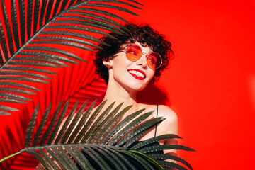 Fashion beauty portrait of young smiling brunette woman with evening stylish  makeup. Sexy model with curly short hair posing in studio. With red bright lips. Tropical palm leaf. In sunglasses