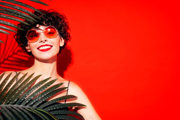 Fashion beauty portrait of young smiling brunette woman with evening stylish  makeup. Sexy model with curly short hair posing in studio. With red bright lips. Tropical palm leaf. In sunglasses