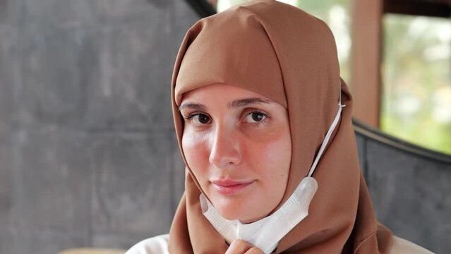 Young woman in a hijab looks directly into the camera, pulls down her face mask. Close portrait shot with blurred background. The lady keeps looking right at us with a little smile on her face