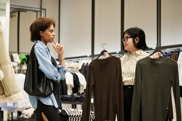Young asian shop assistant offering two jumpers for choice to african american buyer