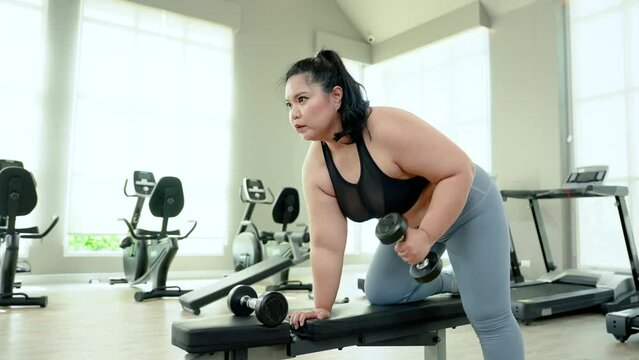 Overweight Asian woman, exercising in gym, exercise by lifting dumbbell with left hand, using right hand support seat, In order tighten muscles each arm, come here regularly every evening.