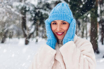 Close up portrait of happy young woman walking under falling snow in winter park wearing white fur coat blue hat mittens