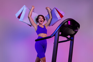 Photo of excited sporty girl using running machine raise shopping bags buy on sport discounts...