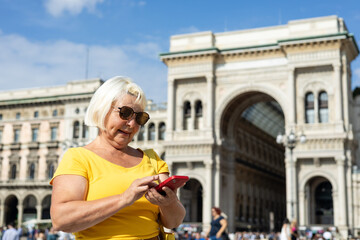 Pretty senior woman staying on the street and holding phone. Tourist checks her smartphone on Duomo...