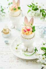 Stuffed or deviled eggs with yolk, shrimp, pea microgreens with paprika in rabbit-shaped stand for easter table decorate fresh cherry or apple blossoms on light background. Traditional dish for Easter