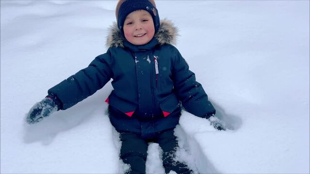A happy boy falls into a snowdrift. Flakes of snow fall on the child's face, he laughs and winces sweetly. Little boy playing in snowdrift and having fun with fresh snow. High quality 4k footage
