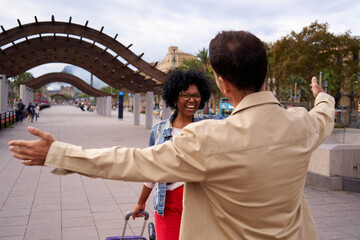 African American woman happily greeting her boyfriend with luggage outdoors. Meeting in the street of two people who have not seen each other for a long time about to hug.