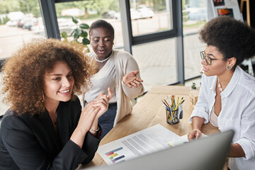 smiling multiethnic businesswomen looking at computer monitor near documents with graphs in office